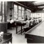 Fila de trabajadoras sentadas en una fábrica pintando manualmente esferas de reloj junto a grandes ventanales