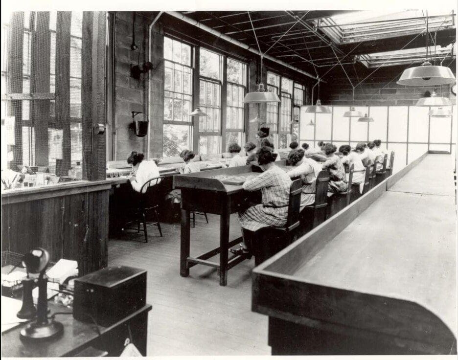 Fila de trabajadoras sentadas en una fábrica pintando manualmente esferas de reloj junto a grandes ventanales