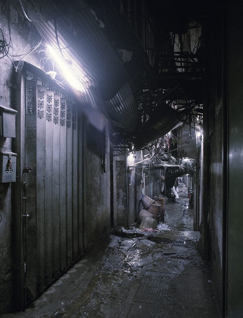 Estrecho callejón interior de la Ciudad Amurallada de Kowloon, apenas iluminado por luces artificiales y cables